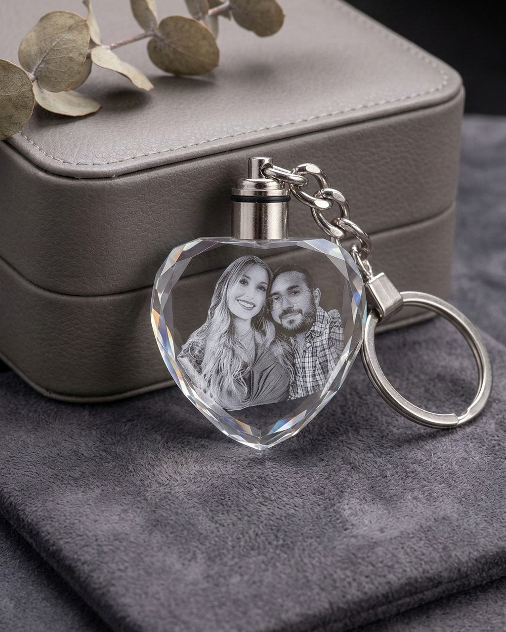 Heart-shaped keychain with engraved photo on a gray surface with a jewelry box in the background.